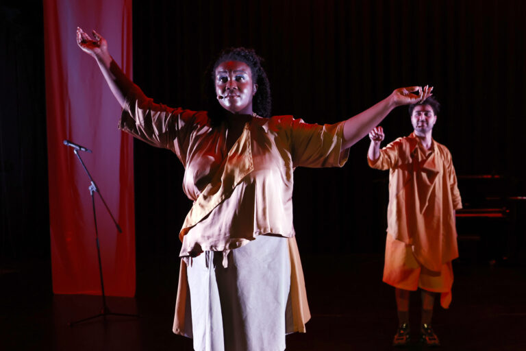 Staande actrice met armen omhoog, met daarachter een acteur die naar haar wijst.