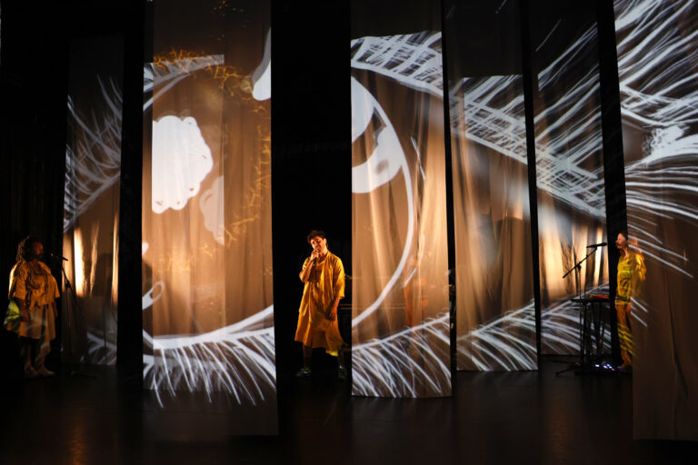 Zingende man en achtergrondkoor op podium tussen wapperende gordijnen.