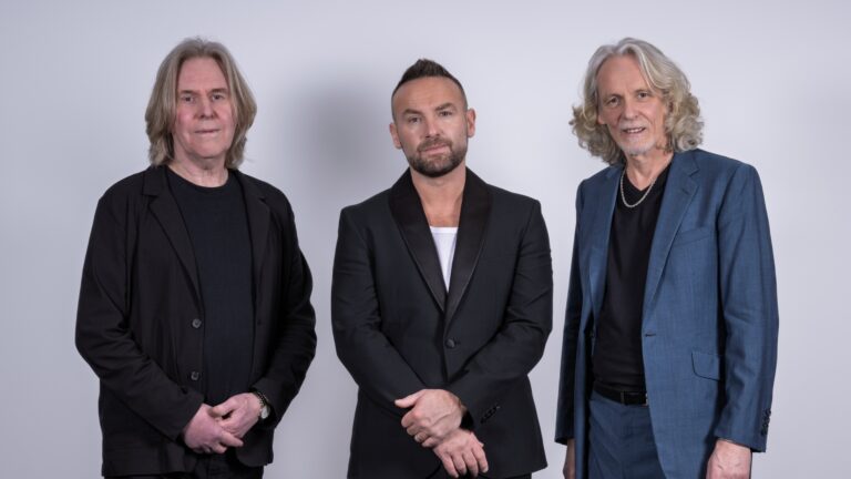 Drie mannen staan naast elkaar voor een lichte achtergrond, gekleed in formele kleding.