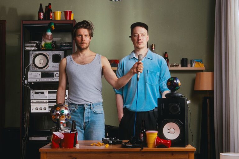 Twee mannen staan in een woonkamer met een muziekstation en een microfoon, omgeven door kleurrijke decoraties.
