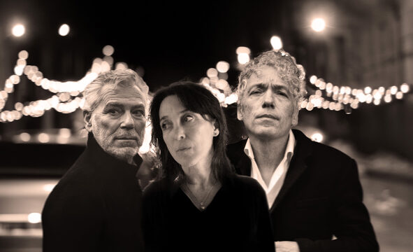 Een vrouw staat tussen twee mannen op straat, met sfeervolle verlichting op de achtergrond.