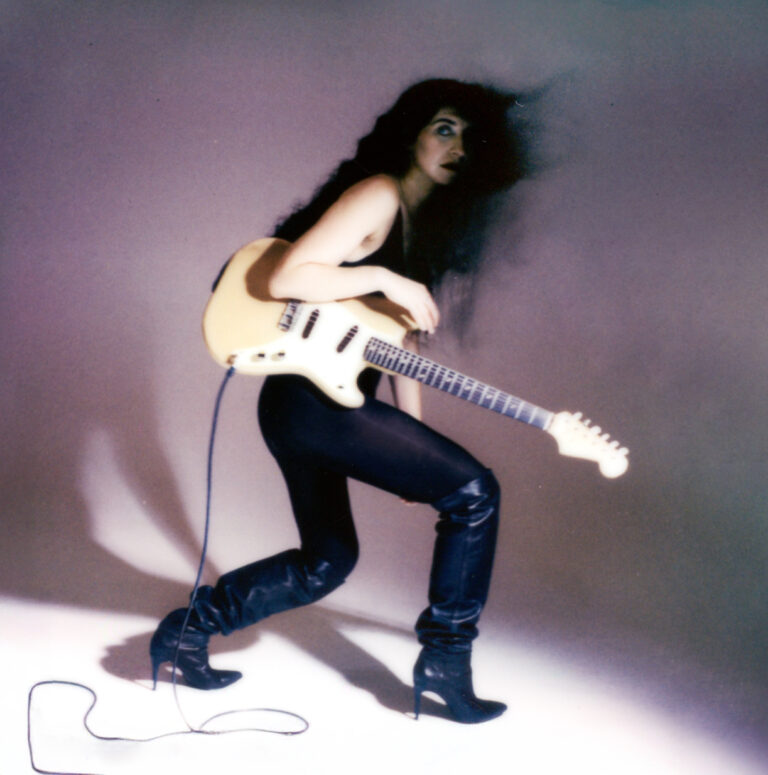 studio portret in jaren '80 stijl van vrouw met lang zwart haar, glimmende leren broek, hoge hakken en een gitaar