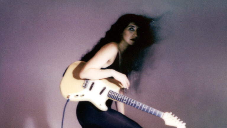 studio portret in jaren '80 stijl van vrouw met lang zwart haar, glimmende leren broek, hoge hakken en een gitaar