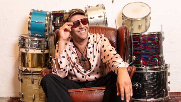 Muzikant met zonnebril en polkadot blouse poseert lachend in stoel tussen drumstellen.