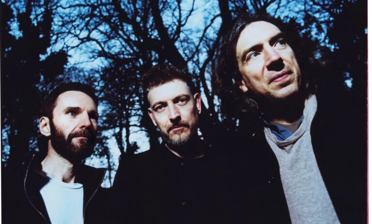 3 leden Snow Patrol staan buiten met bomen op achtergrond.