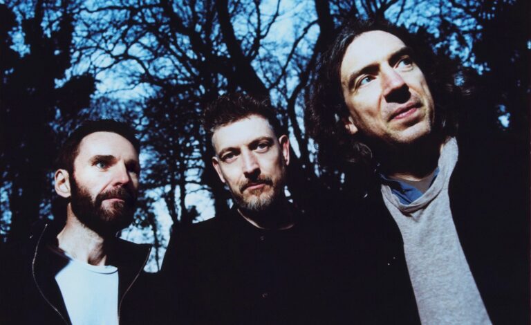 3 leden Snow Patrol staan buiten met bomen op achtergrond.