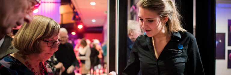 Een vrouw kijkt door een glazen deur, terwijl mensen in een fel verlichte ruimte op de achtergrond zichtbaar zijn.