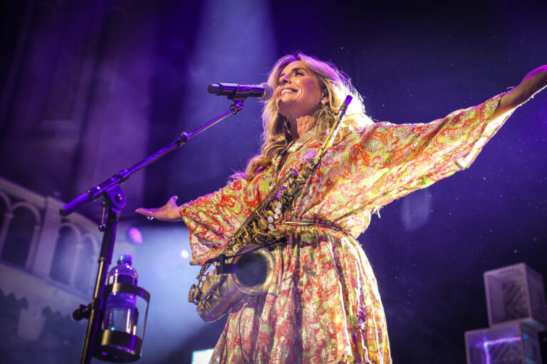 Blonde vrouw staat op het podium met beide armen uitgestrekt en een glimlach voor de microfoon.