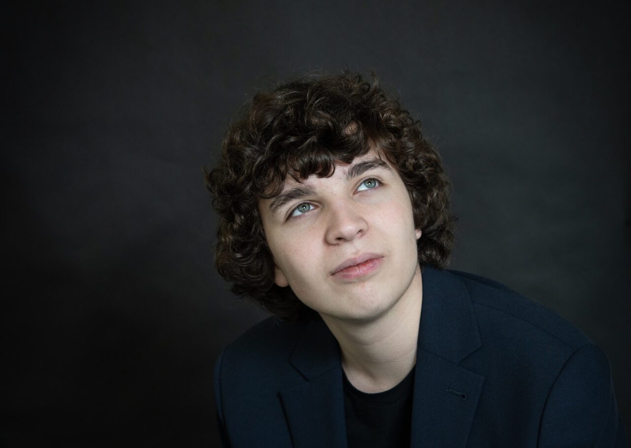 Portret van een persoon met donker krullend haar tegen een donkere achtergrond, kijkend naar boven-links.