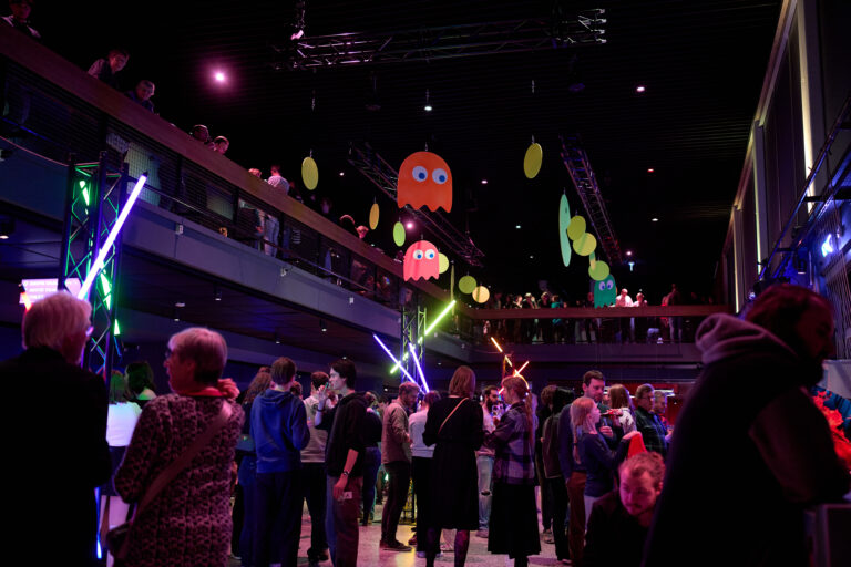 Een drukbezochte zaal waarin mensen dansen tijdens een evenement, verlicht door felgekleurde neonlichten en glow-in-the-dark decoraties.