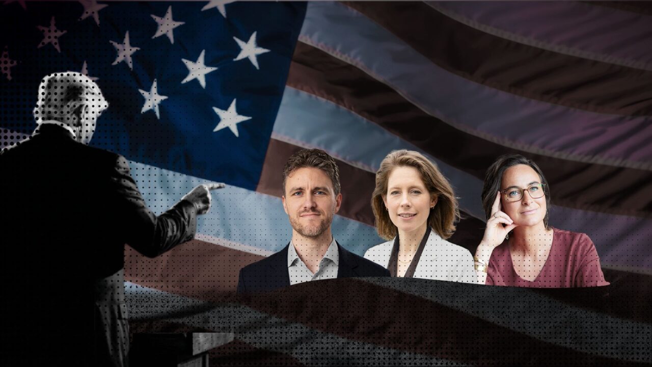 Drie mensen in formele kleding staan op een podium met een grote Amerikaanse vlag op de achtergrond.