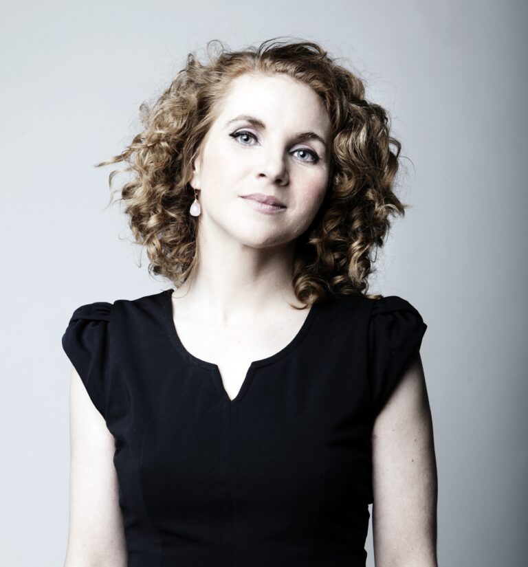 Portret van een vrouw met krullig lichtbruin haar en een zwarte blouse tegen een lichte achtergrond.
