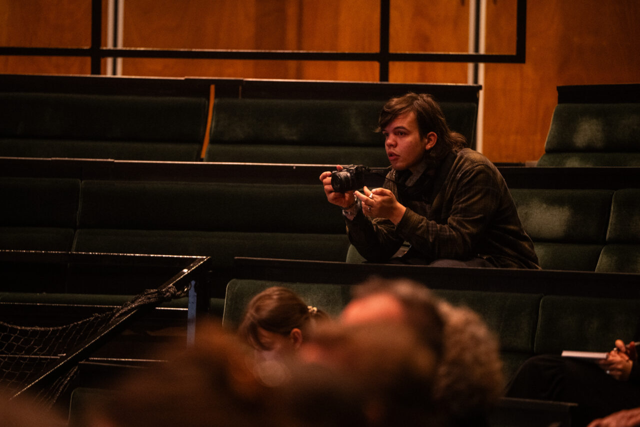 Een man in donkere kleding houdt een camera voor zijn gezicht terwijl hij in een theaterzaal zit.