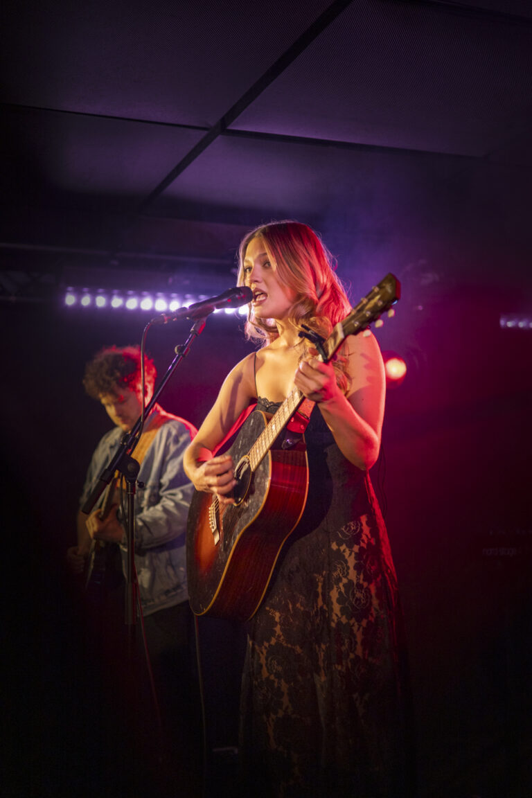 Een zangeres speelt gitaar en zingt op een podium onder paars en roze licht, met een tweede muzikant op de achtergrond.