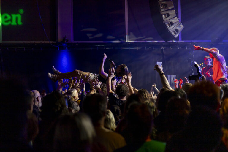 Een volle concertzaal met een menigte die hun armen omhoog steekt onder blauw-paarse podiumverlichting.