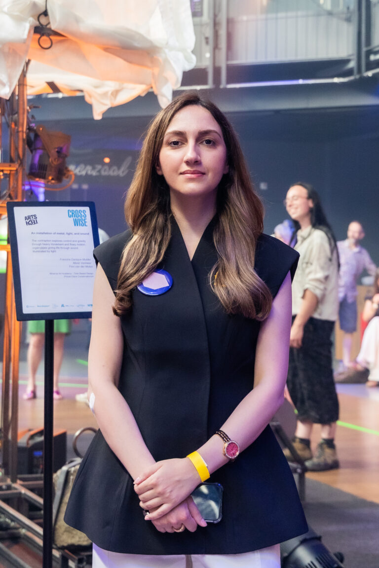 Een jonge vrouw met lang bruin haar draagt een donker jurkje en een badge op haar borst op een druk evenement.