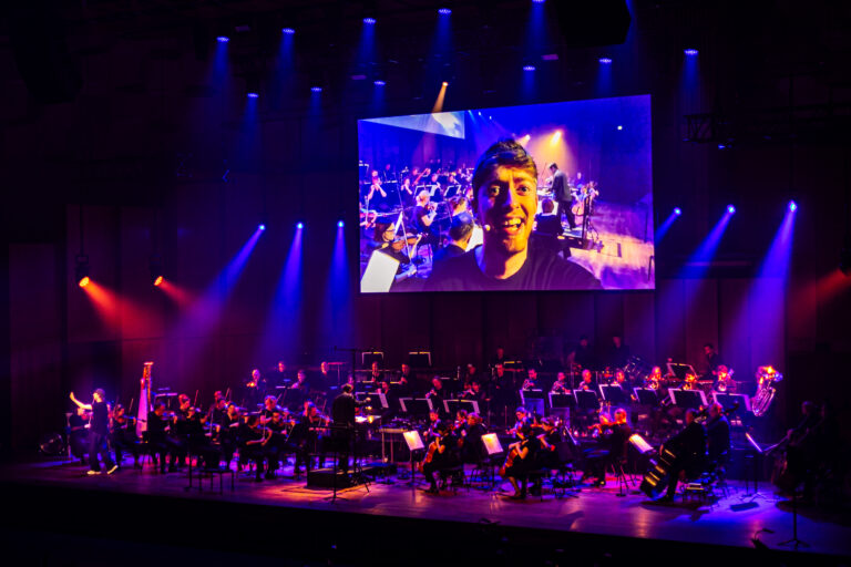 Podium in een concertzaal met blauwe verlichting en een groot scherm dat een close-up van de performer toont terwijl het publiek kijkt.