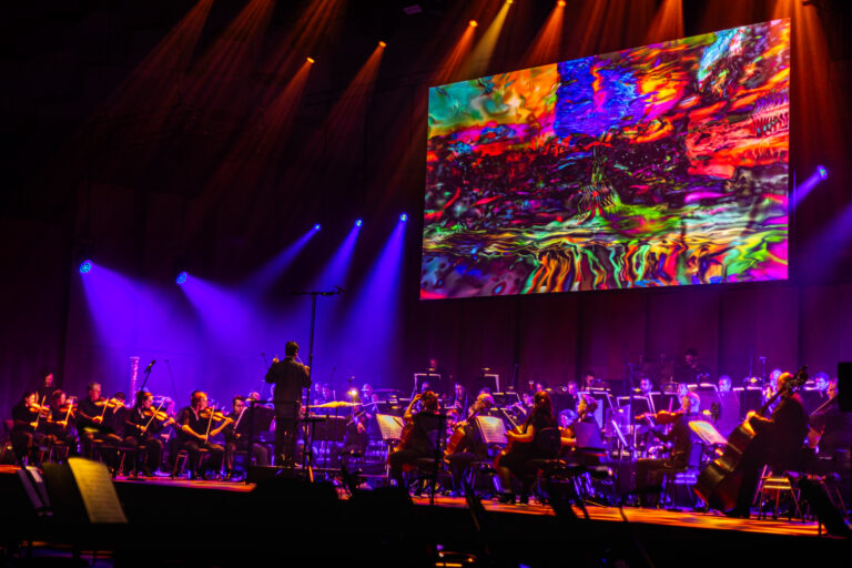 Een orkest speelt op een fel verlicht podium, met een groot kleurrijk scherm boven hen.