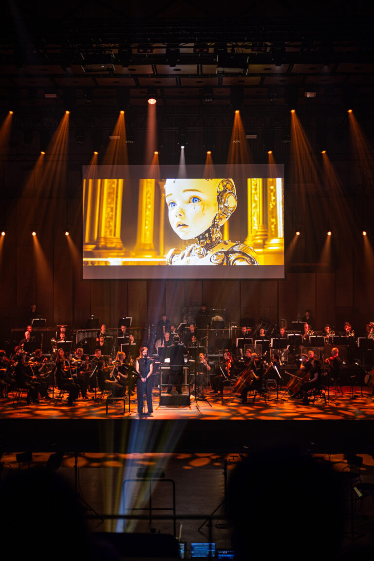 Op een podium met een orkest is een groot scherm te zien dat een close-up van een gezicht toont.