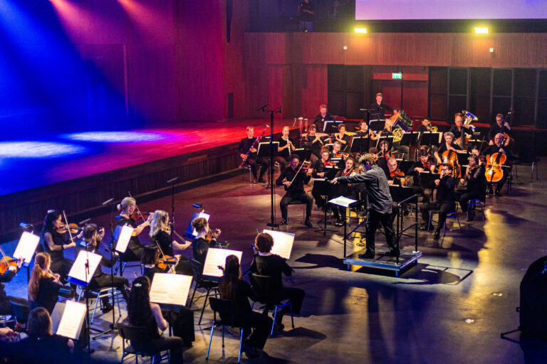Een orkest onder leiding van een dirigent speelt op een podium in een concertzaal.