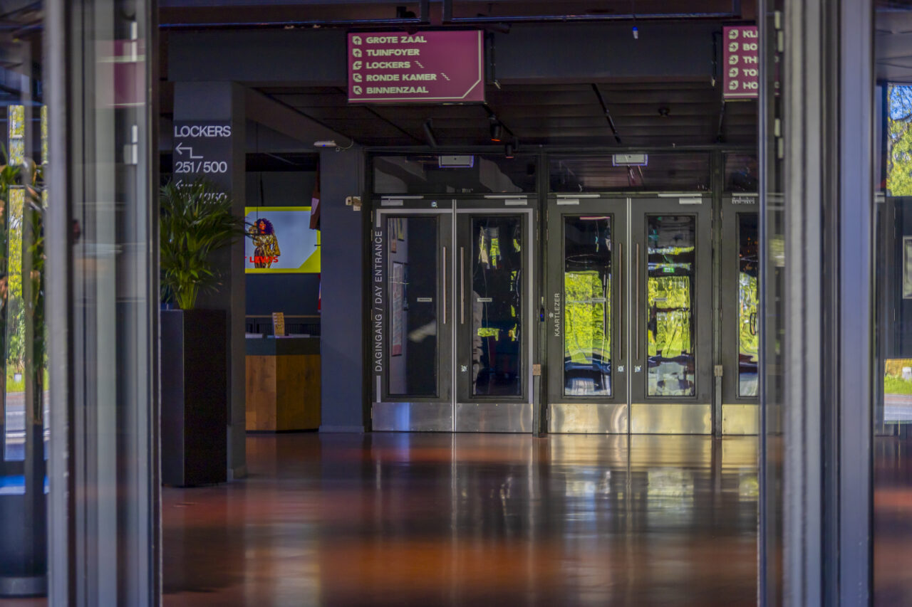 Glazen deuren met daarboven routing bordjes daarnaast zie je nog een narrowcasting scherm
