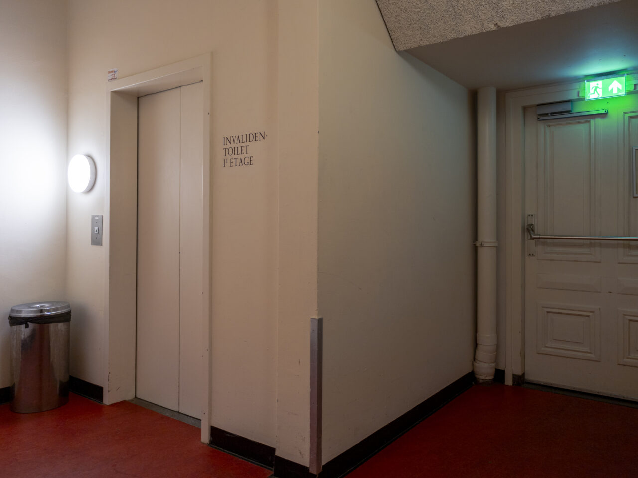 Een grote nooduitgang deur met daarnaast een witte liftdeur met rode vloerbedekking.